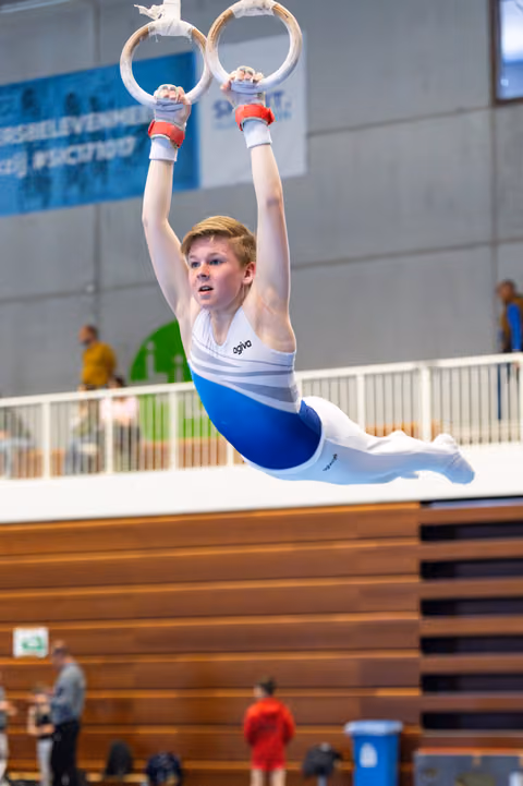 Young athlete performing a strong hold position on still rings during indoor competition, displaying intense focus and strength