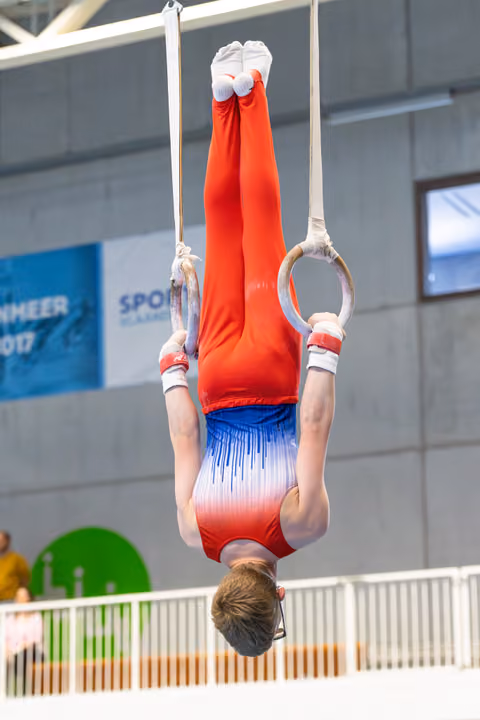 Athlete performing inverted hang on still rings with extended legs and pointed toes during indoor competition