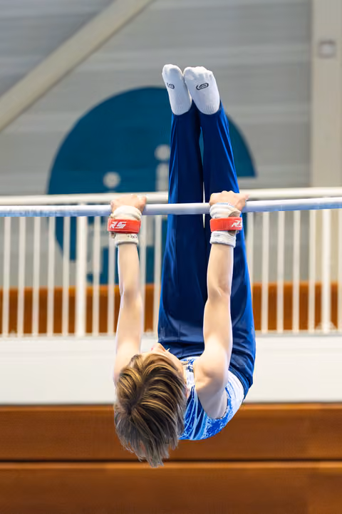 Young athlete hanging upside down from horizontal bar, legs extended upward, wearing blue uniform and white grips