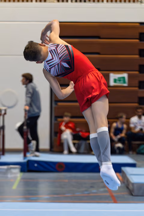Athlete performs dynamic backbend jump in red uniform and patterned leotard during floor routine at indoor competition