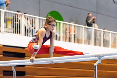 Young athlete in burgundy leotard and red pants performs on parallel bars while spectators watch from balcony above