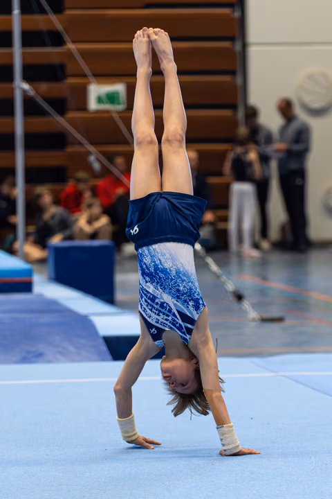 Young athlete performs a vertical handstand during floor exercise, wearing blue leotard with white support wraps on wrists