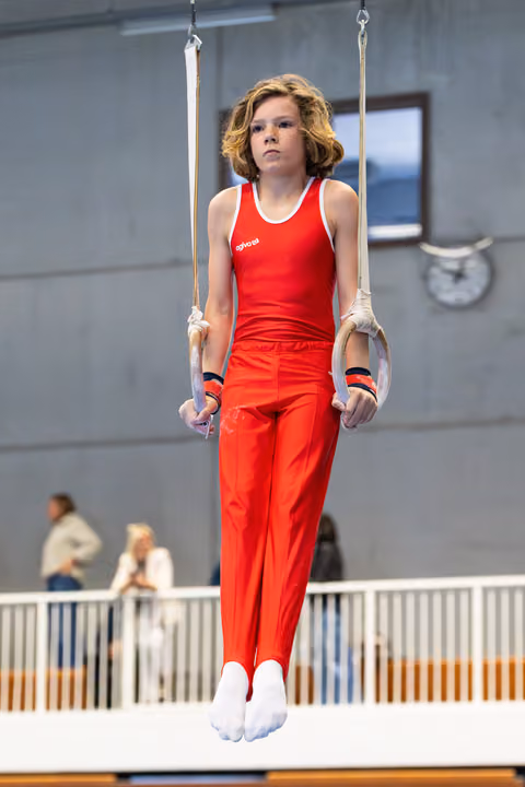 Young gymnast in red uniform performs a hold position on still rings during competition at indoor venue