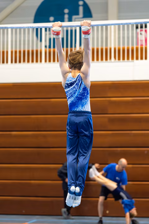 Young athlete in blue leotard hangs from high bar with arms extended overhead during indoor training session