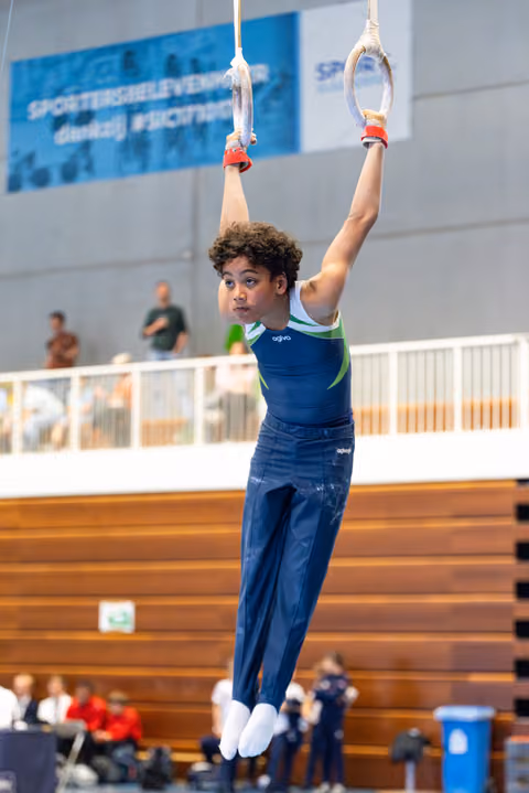 Young athlete hanging from rings with focused expression during routine, wooden floor and spectators visible in background