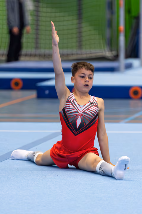 Young gymnast performs floor routine in seated split position with raised arm, wearing red and striped leotard