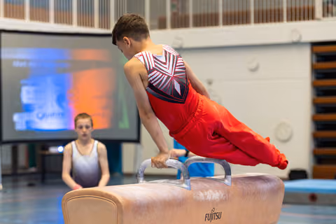 Athlete performs horizontal planche hold on pommel horse apparatus during training session, teammate observing in background