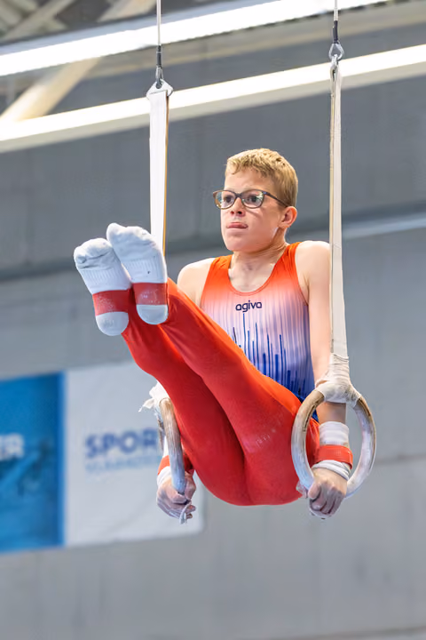 Young athlete performing on still rings with focused expression, wearing glasses and an orange-to-blue gradient leotard