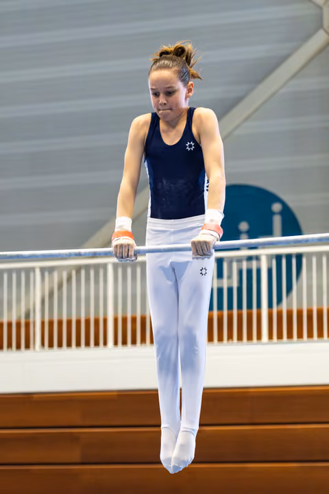 Young gymnast in navy leotard and white pants performs on uneven bars, displaying focused concentration during her routine