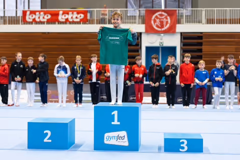 Young athlete on first place podium holding up a green team shirt while fellow competitors with medals stand behind