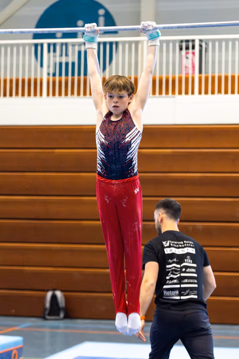 Young athlete hangs from horizontal bar with focused expression while coach spots from below in indoor training facility