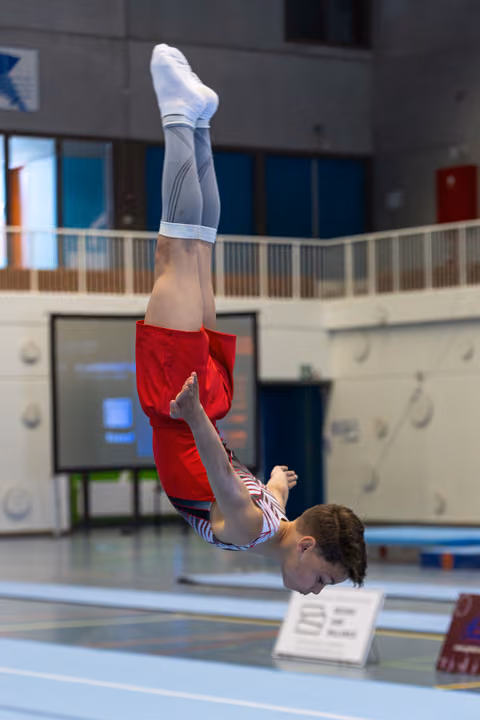 Athlete performs inverted position during floor routine, body fully extended in red and white striped uniform at indoor sports venue