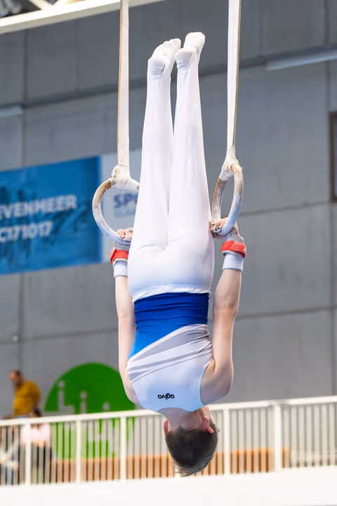 Athlete performing inverted hang on rings with straight body position, wearing white and blue uniform at indoor venue
