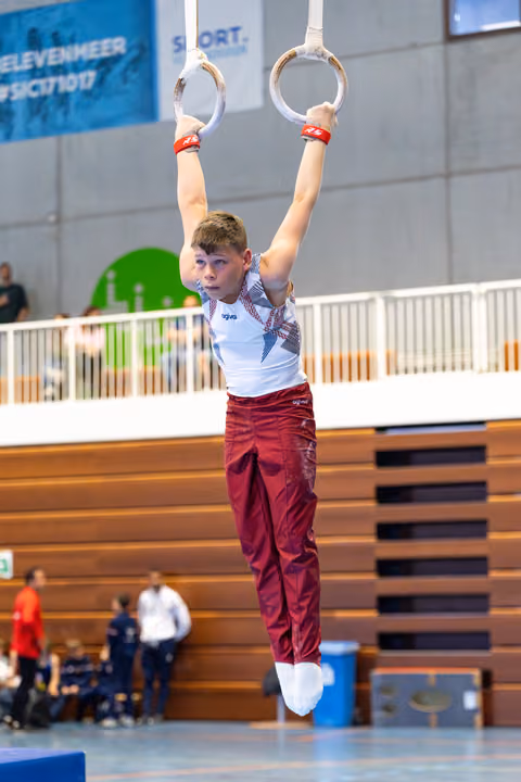 Young athlete hangs from still rings with focused expression, wearing white and maroon uniform during indoor competition