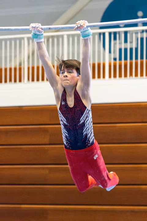 Young athlete hanging from horizontal bar with intense concentration, wearing purple leotard and red shorts in gymnasium