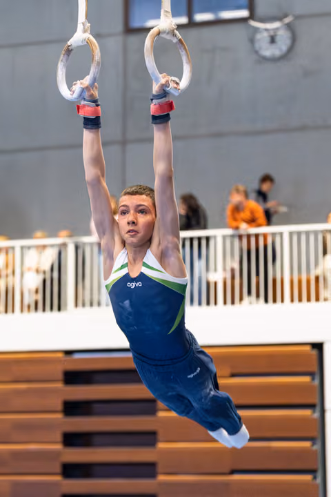 Young athlete hanging from still rings with arms extended overhead during routine, wearing navy blue leotard in indoor venue
