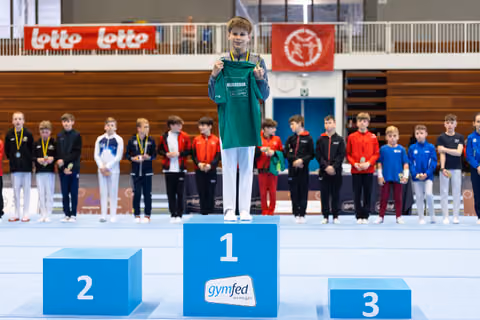 Young athlete in green jacket stands on first place podium holding gold medal, with competitors lined up behind in gymnasium