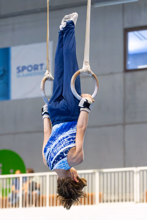 Young athlete performing an inverted hang on still rings, legs extended upward, displaying strength and control in indoor gym