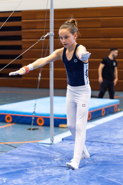 Young gymnast in navy leotard and white pants stands focused on the mat, arm extended in preparation for her routine