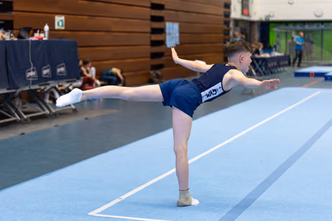 Athlete performs a horizontal scale balance during floor routine, arms extended gracefully on blue spring floor