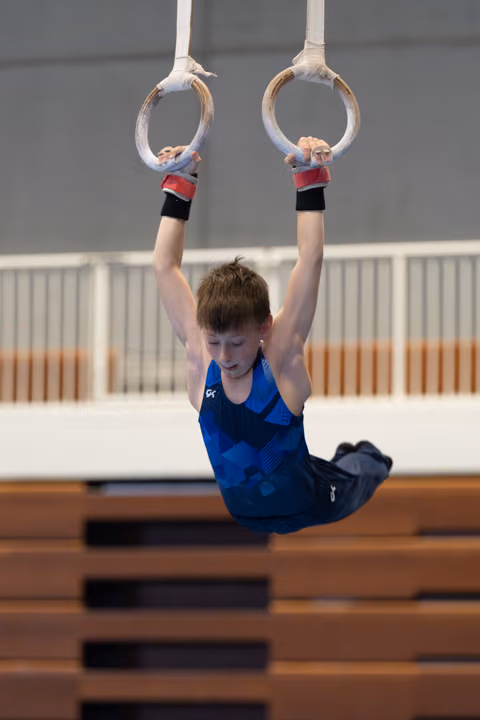Young gymnast suspended from rings with bent knees, wearing blue leotard and wrist guards in indoor training facility
