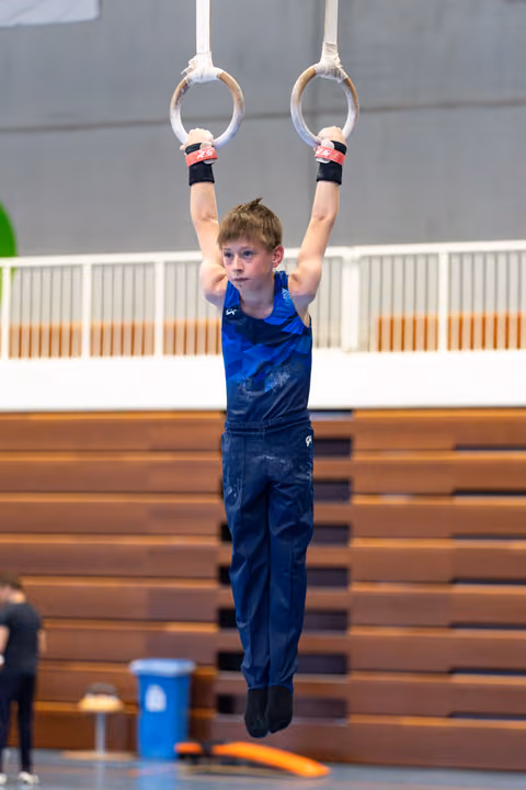 Young athlete in blue tank top hanging from still rings with focused expression during training at indoor gymnasium