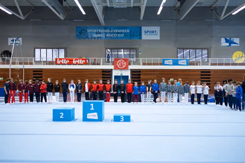 Teams lined up behind medal podium at indoor sports hall, athletes wearing coordinated team tracksuits in red, black, blue, and grey
