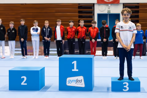 Bronze medalist stands proudly on third place podium wearing white tracksuit while other competitors line up in background