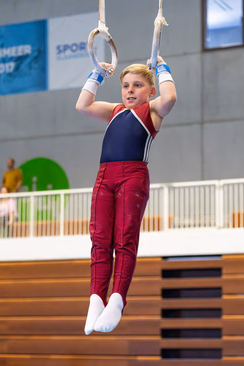 Young athlete hanging from rings in red and navy uniform, displaying focus during training at indoor sports facility