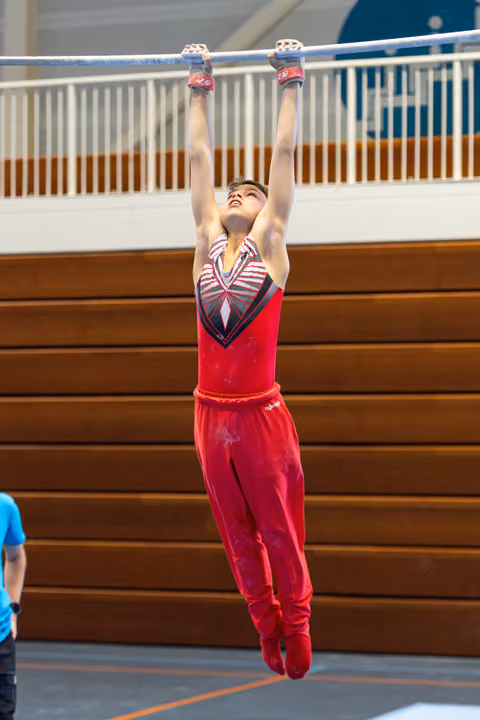 Young athlete in red uniform hangs from high bar with arms fully extended, looking up during training in indoor gymnasium
