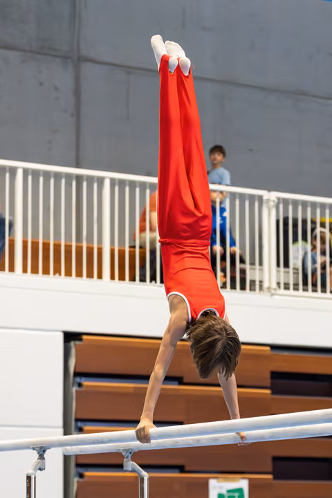 Athlete performing a vertical handstand on parallel bars wearing red uniform, demonstrating balance and strength