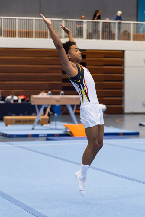 Athlete performing floor routine with arms raised during jump, white leotard with colorful design, indoor gymnasium