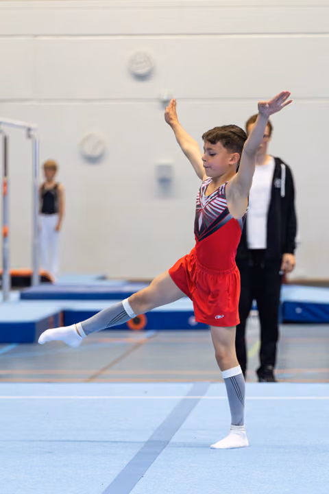 Young athlete performs arabesque on floor exercise, wearing red shorts and grey knee socks, with coach observing in background