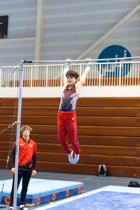 Young athlete hanging from horizontal bar with arms extended, wearing red pants and gradient leotard, coach watching below