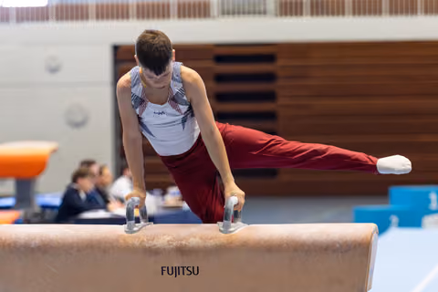 Gymnast performs pommel horse routine with extended legs, wearing maroon pants and white tank top in competition hall