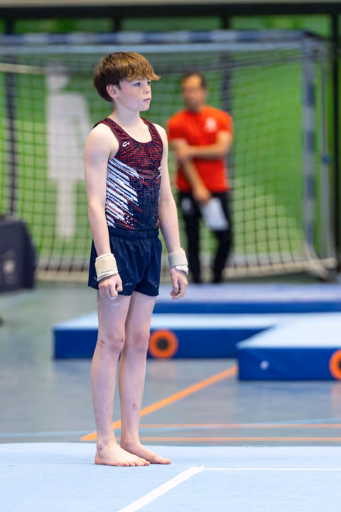 Young gymnast stands at attention on floor mat, focused expression, wearing sequined leotard with wrist supports