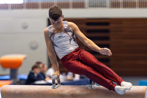 Young athlete performing a pommel horse routine with focused concentration, wearing white and red uniform at indoor competition