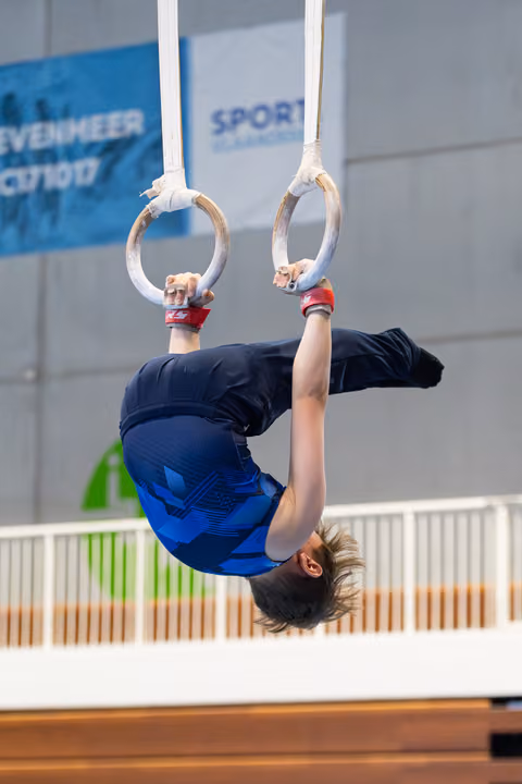 Athlete performing an inverted pike position on still rings, wearing red grips and blue shorts in indoor sports hall