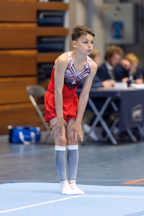 Young athlete in red shorts and striped leotard stands ready on floor mat, hands on knees, displaying focused concentration