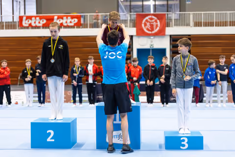 Three young athletes stand on award podium wearing medals, with other participants watching in the background at indoor sports hall