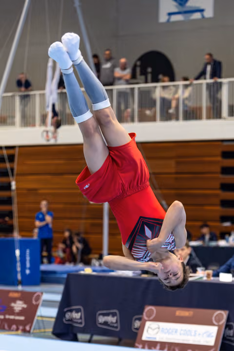 Female gymnast performs inverted handstand on floor exercise mat during competition, legs extended vertically in red leotard