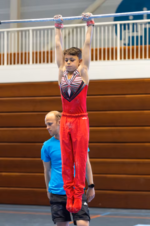 Young athlete in red outfit hanging from horizontal bar with arms extended while coach in blue shirt spots from below