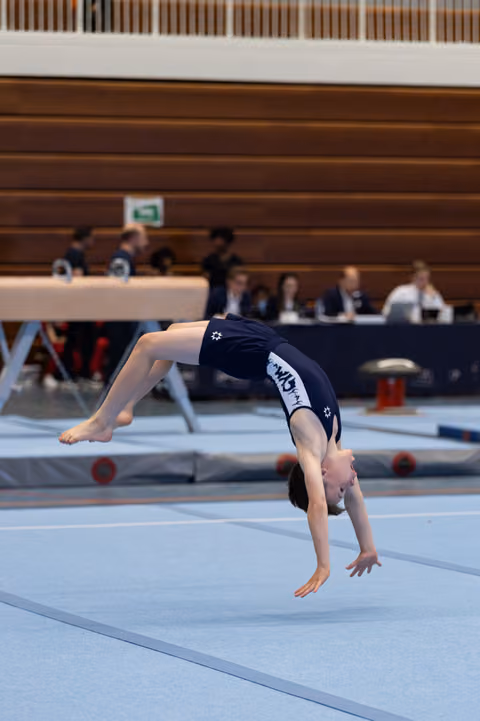 Gymnast performing a backward walkover on floor exercise mat during competition, judges seated in background