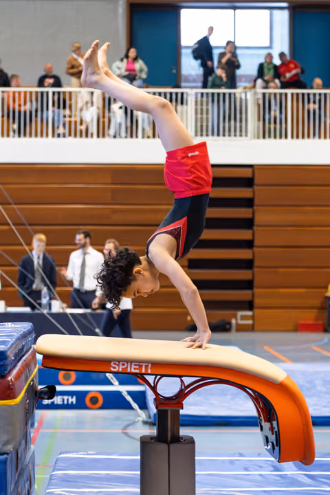 Female gymnast executes a handstand on vault apparatus during competition, displaying strength and balance with judges watching