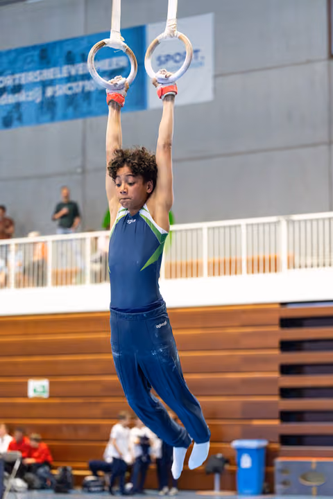 Young athlete grips still rings during routine, body fully extended with focused expression in indoor sports hall