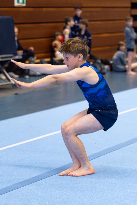 Young athlete performs floor exercise in blue leotard, focused expression in deep squat with arms extended forward
