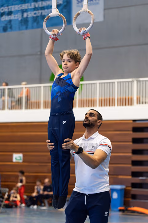Young gymnast hanging from rings with focused expression while coach stands below providing support during training