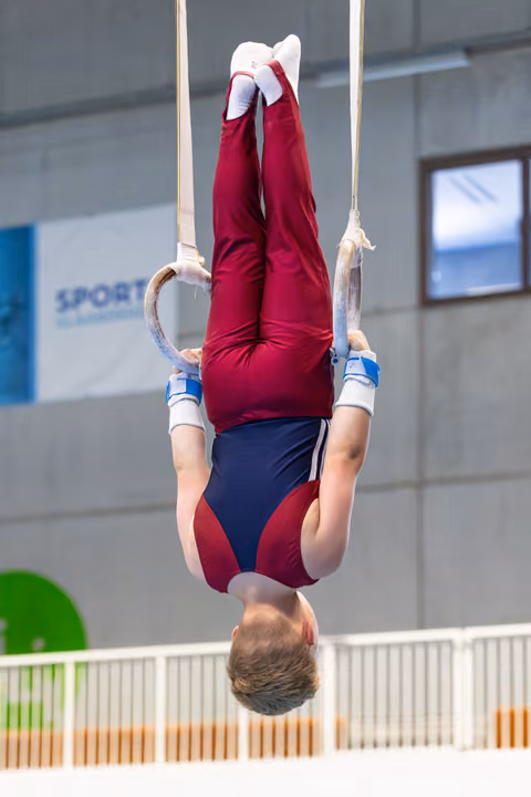 Young athlete performs an inverted hang on rings during routine, displaying strength and control in maroon and navy uniform