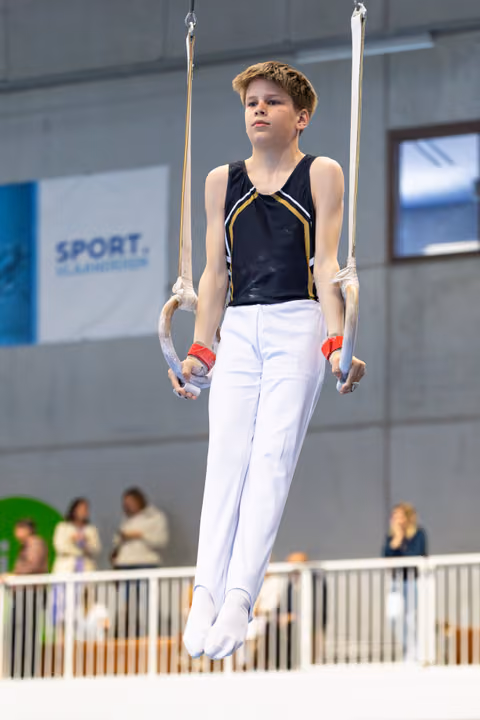 Young athlete suspended on still rings, displaying concentration and controlled form during routine at indoor sporting venue