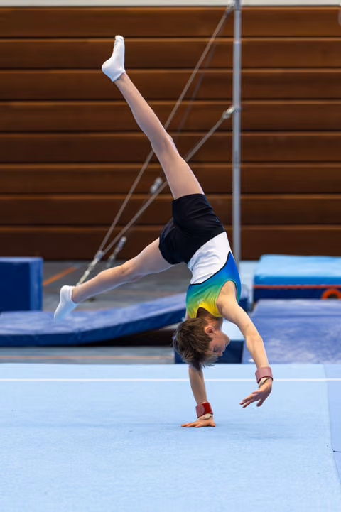Young gymnast executing a one-handed handstand on floor exercise mat, demonstrating strength and balance in training facility